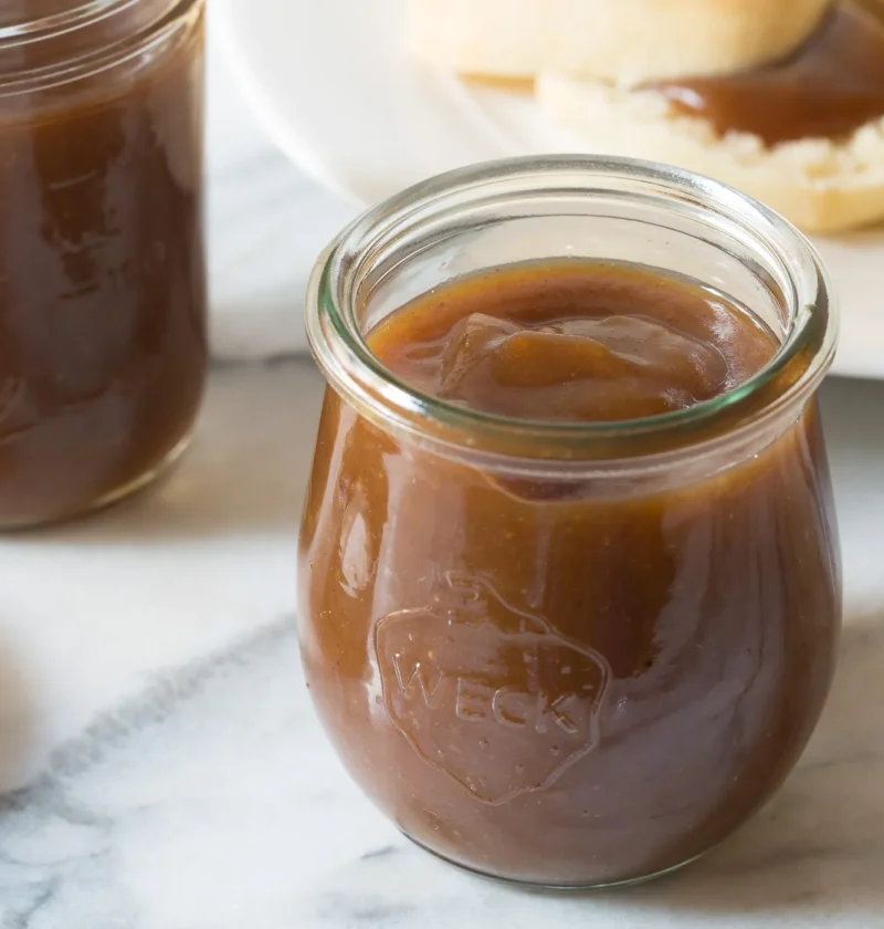 A jar of homemade pioneer woman apple butter with a thick, dark brown spread next to fresh apples
