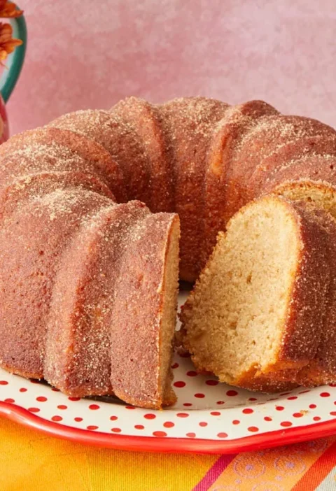 A golden apple cider donut cake on a wire rack coated in cinnamon sugar with a slice removed showing the tender crumb