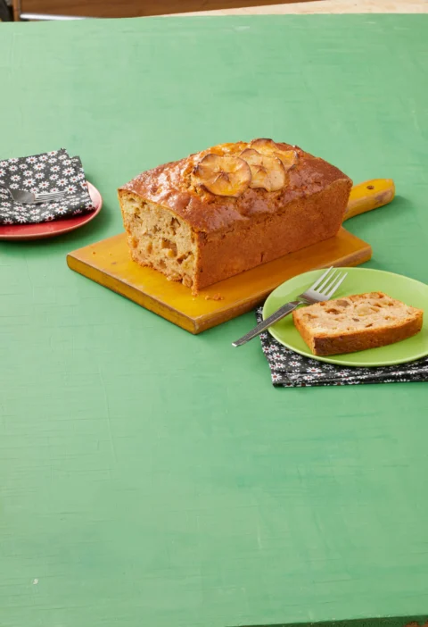 A golden loaf of pioneer woman apple cinnamon bread with thin apple slices on top and an apricot jam glaze, sliced on a wooden cutting board