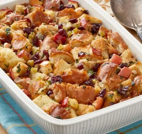 A baking dish filled with golden brown pioneer woman apple cranberry stuffing topped with crispy bread cubes, dried cranberries and chopped apples