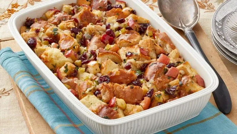 A baking dish filled with golden brown pioneer woman apple cranberry stuffing topped with crispy bread cubes, dried cranberries and chopped apples