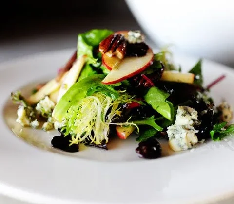 Apple pecan and blue cheese salad with dried cherries in a large bowl, topped with thin apple slices, pecan halves and crumbled blue cheese over spring greens
