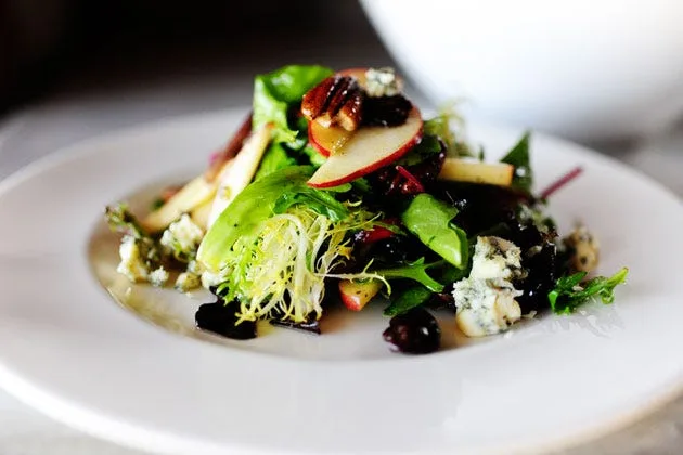 Apple pecan and blue cheese salad with dried cherries in a large bowl, topped with thin apple slices, pecan halves and crumbled blue cheese over spring greens