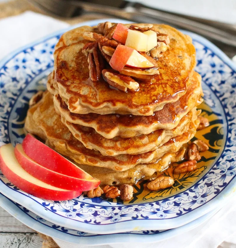 A stack of golden apple spiced pancakes topped with fresh fruit and maple syrup on a white plate