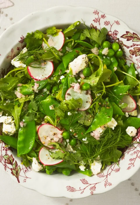 A bowl of arugula and three pea salad with crumbled goat cheese, fresh herbs and sliced radishes
