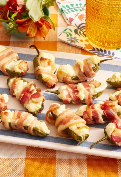 Bacon-wrapped jalapeno thingies lined up on a baking rack, each stuffed with cream cheese and wrapped in crispy bacon