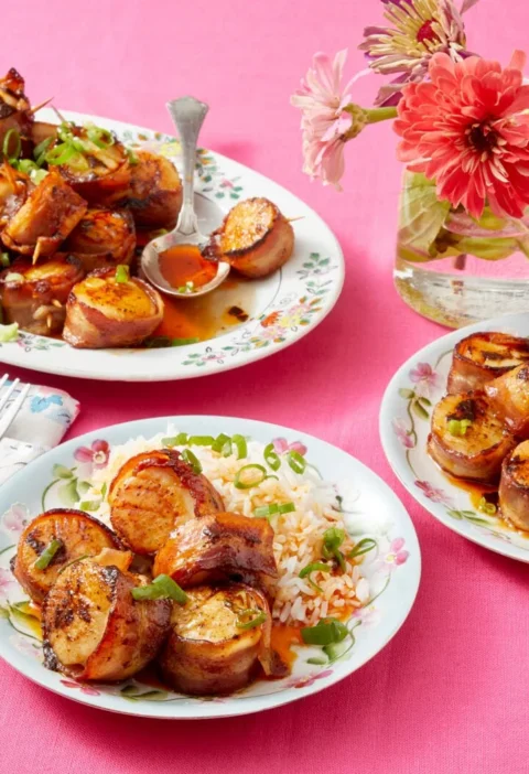 Bacon wrapped scallops with chili butter drizzled on top, arranged on a serving plate with wooden skewers