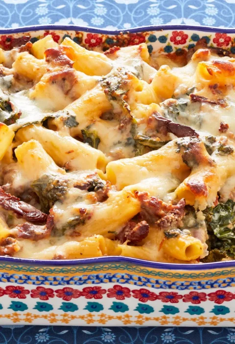 Baked pasta with Italian sausage, kale and melted mozzarella in a white baking dish, showing golden bubbly cheese on top
